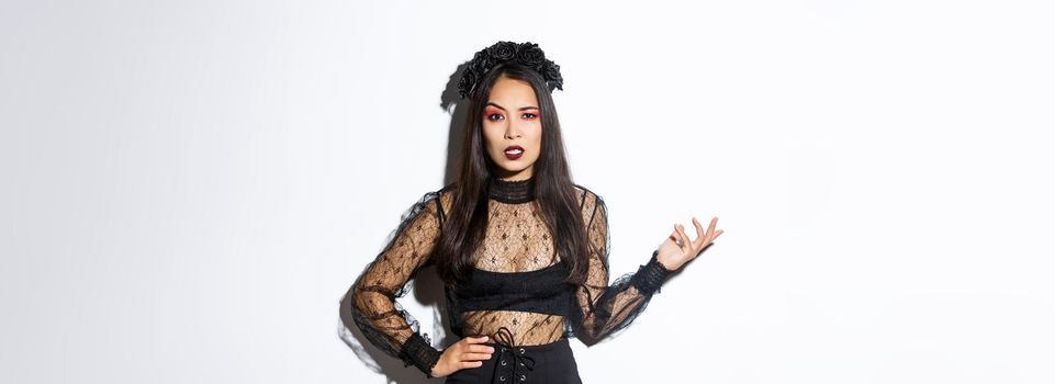 Angry and confused asian woman in witch costume looking bothered by annoying quetion, raising hand up and stare at camera pissed-off. Female at halloween party standing frustrated.