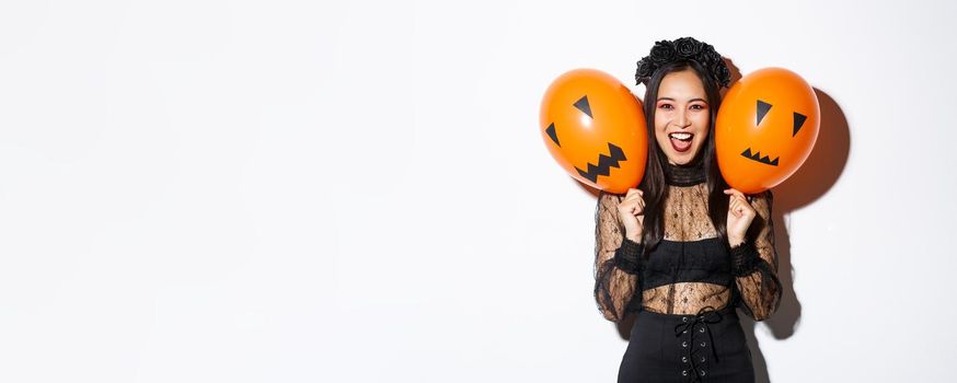 Image of asian girl in evil witch costume holding two orange balloons with scary faces, celebrating halloween, standing over white background.