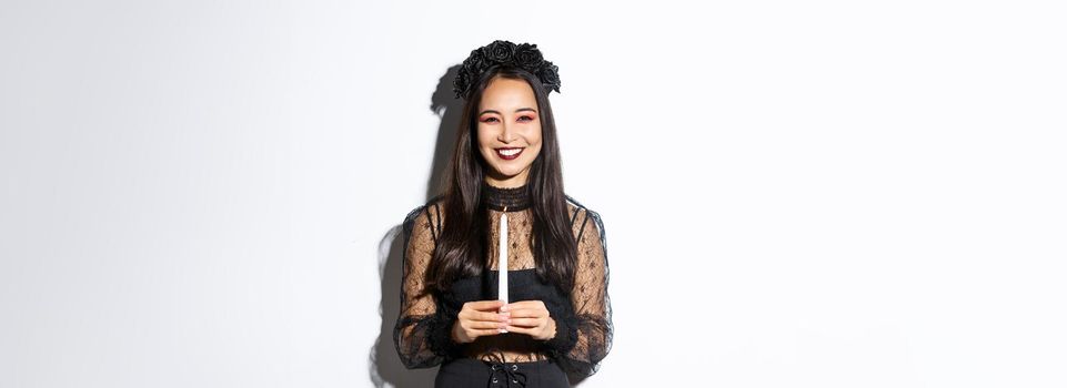 Beautiful asian woman having fun on halloween party, dressed-up as witch with candle, standing over white background.
