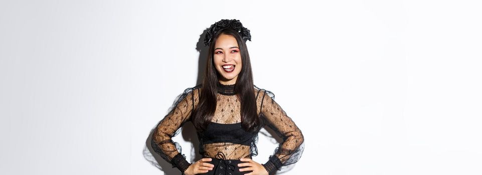 Image of carefree smiling asian woman in gothic dress and black wreath looking thoughtful at upper left corner, licking lips from desire or temptation, standing over white background.