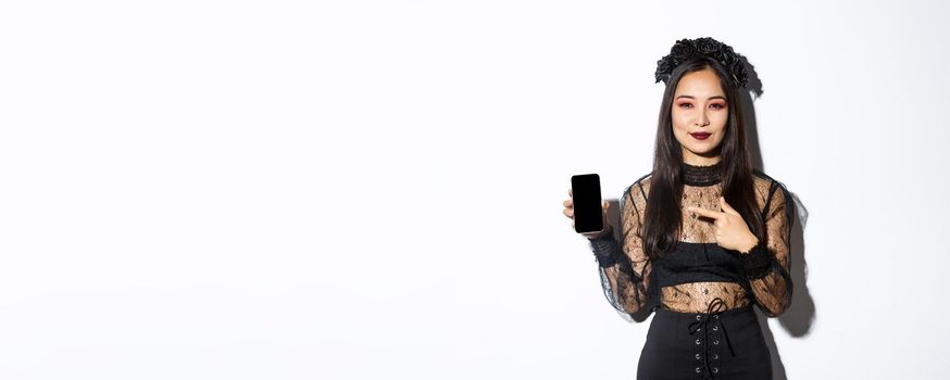 Young elegant woman in gothic dress and black wreath pointng finger at smartphone screen with pleased smile on her face, standing over white background.