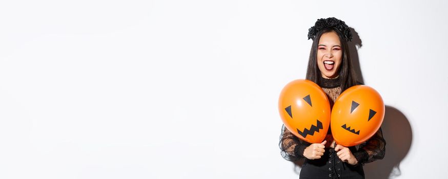 Image of asian girl in evil witch costume holding two orange balloons with scary faces, celebrating halloween, standing over white background.