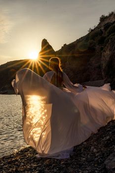 A mysterious female silhouette with long braids stands on the sea beach with mountain views, Sunset rays shine on a woman. Throws up a long white dress, a divine sunset