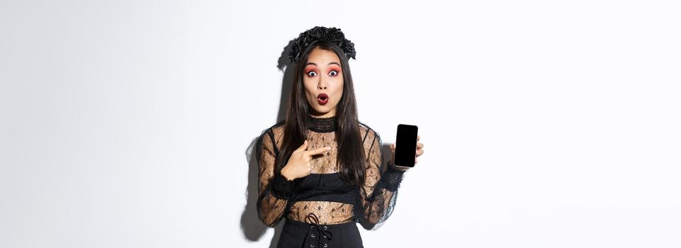 Surprised asian girl in black gothic dress with wreath, gasping amused and pointing finger at mobile phone display, showing halloween banner or promo, standing over white background.