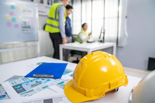 Concept of engineering equipments of protective helmet for competent engineer on piles of paper works. Blurred image of engineers and employer working together at the desk.