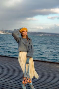 Outdoors fashion portrait of a beautiful middle aged woman walking on the beach. Marine background. Dressed in a stylish warm blue sweater, yellow skirt and beret