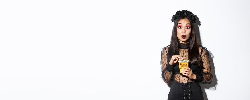 Image of amazed asian woman in witch halloween costume looking at camera while holding sweets in trick or treat bag, standing over white background.