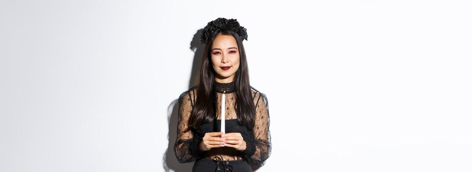 Image of beautiful woman celebrating halloween in witch costume, holding candle and squinting at camera suspicious, standing over white background.