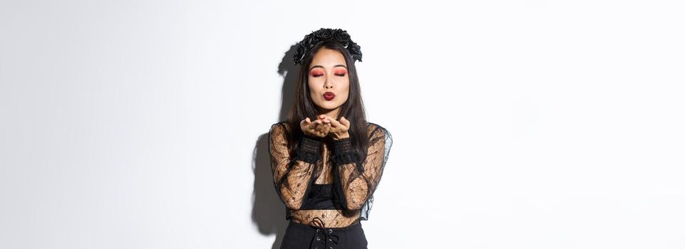 Image of beautiful and elegant asian woman in witch costume, close eyes and sending air kiss t camera, wearing gothic dress with black wreath, standing over white background.