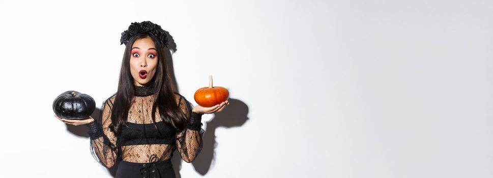 Portrait of surprised asian girl in witch costume holding two pumpkins and gasping amazed while looking at camera, standing over white background.