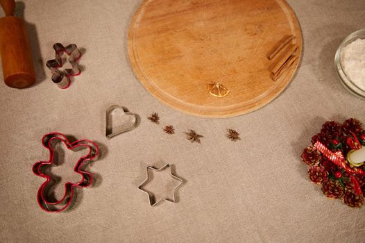 Overhead view of wooden cutting board, rolling pin, various cookie cutters for Christmas gingerbread pastries, star anise seasoning on a linen tablecloth in the home kitchen. Concept of festive baking