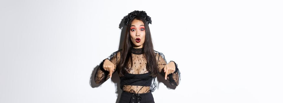 Impressed asian woman in evil witch costume pointing fingers down, showing halloween banner, making announcement. Girl wearing black lace dress and wreath, standing white background.