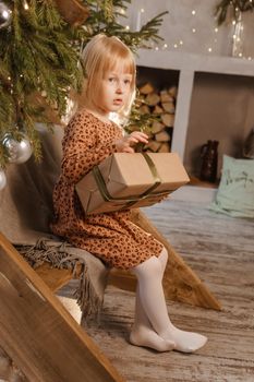 A little blonde girl is sitting on a wooden staircase in a Scandinavian interior decorated in a New Year's style. A child holds a Christmas gift in a craft package.