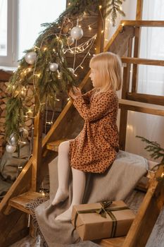 A little blonde girl is sitting on a wooden staircase in a Scandinavian interior decorated in a New Year's style. A child holds a Christmas gift in a craft package.