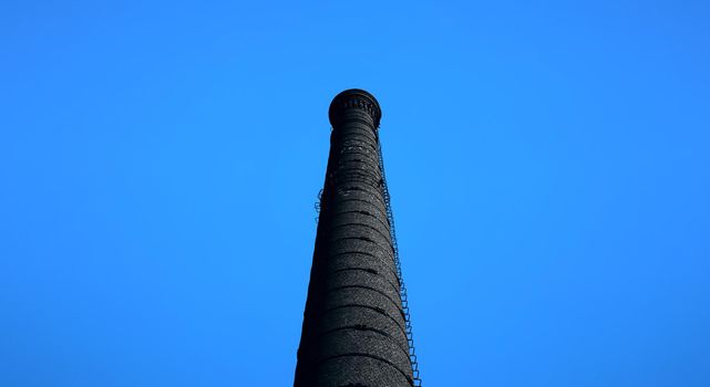 A large chimney in an old factory. smoke stack An old black brick chimney against a blue sky.