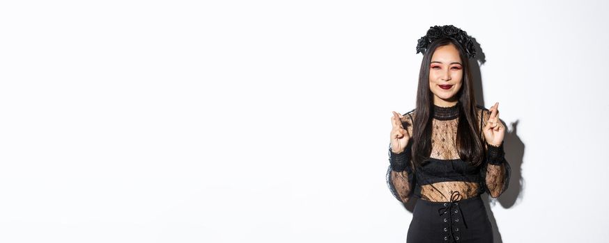 Image of happy asian girl who loves celebrating halloween dressed-up as witch, smiling hopeful and cross fingers for good luck, making wish, standing over white background.