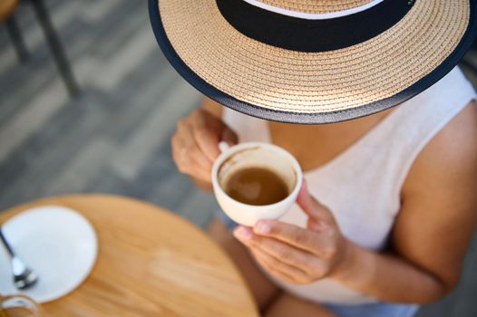 View from above of an unrecognizable young woman in straw hat and summer casual wear, holding a white cup of coffee americano, enjoying her coffee break outdoor, on a cozy summer terrace