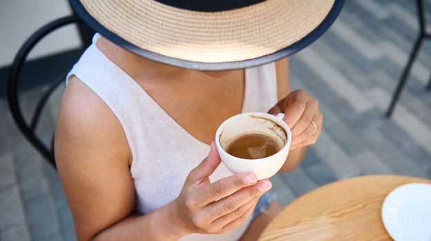 Overhead view of an unrecognizable mysterious woman in straw hat and summer casual dress, holding a white cup of coffee americano, enjoying her coffee break outdoor, on a cozy summer terrace