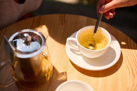 Top view of a woman's hand using a tea spoon, stirring sugar in the white teacup with healthy antioxidant herbal hot drin. Detox therapy. Tea time. Selective focus