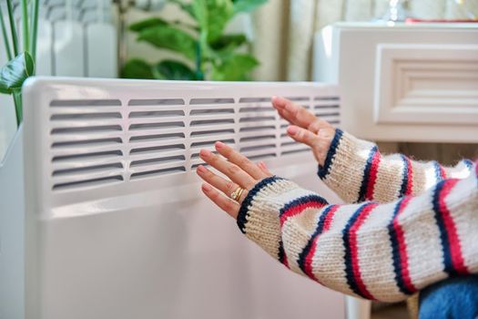 Close-up of a woman's hands warming near an electric heating radiator. Heating cold season autumn winter, comfort, energy saving, crisis, home lifestyle concept