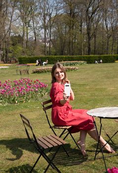 A young beautiful girl in a red dress in a spring park plays with her hair, flirts and takes a selfie on a mobile phone, tulips bloom and people walk in the park, pamper themselves.High quality photo