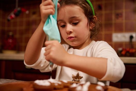 Adorable little girl, lovely daughter standing at a kitchen table, squeezing baking bag with culinary glaze and decorating gingerbread cookies with icing, in Christmas home atmosphere. December 25