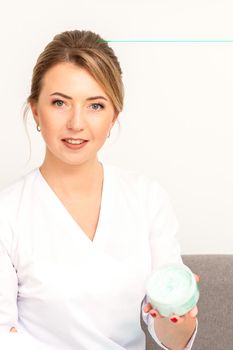 Close-up portrait of young smiling female caucasian healthcare worker standing with jar of cream beauty product on white background.