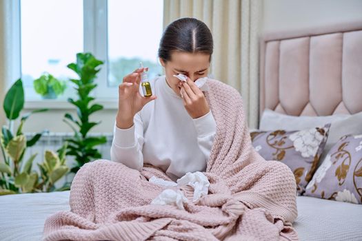 Young teenage female sitting in bed with runny nose medicine spray drops, sneezing into tissues, burying her nose. Autumn winter, rhinitis runny nose stuffy nose, treatment, flu season