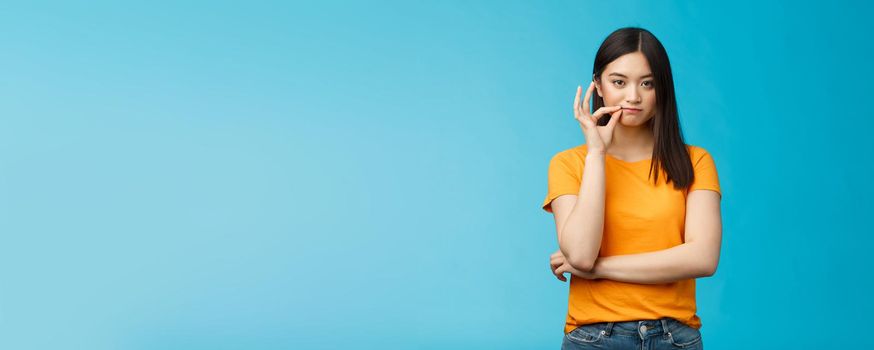 Serious-looking asian female friend promise keep secret, seal lips, hold zip near mouth look focused determined, stay silent and speechless, stand wearing yellow t-shirt blue background.