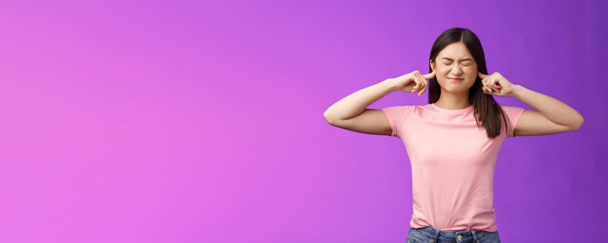 Displeased annoyed asian brunette woman fed up hearing loud parents yell, close eyes grimacing, plug ears fingers, disturbed bothering sound, stand purple background unsatisfied. Copy space