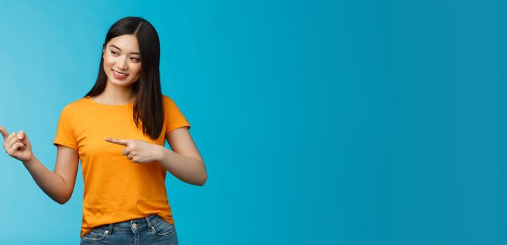 Tender lovely asian girl with dark haircut look curiously pointing left, interested in new product in store, stand blue background amused, entertained, observe exhibition, wear yellow t-shirt.