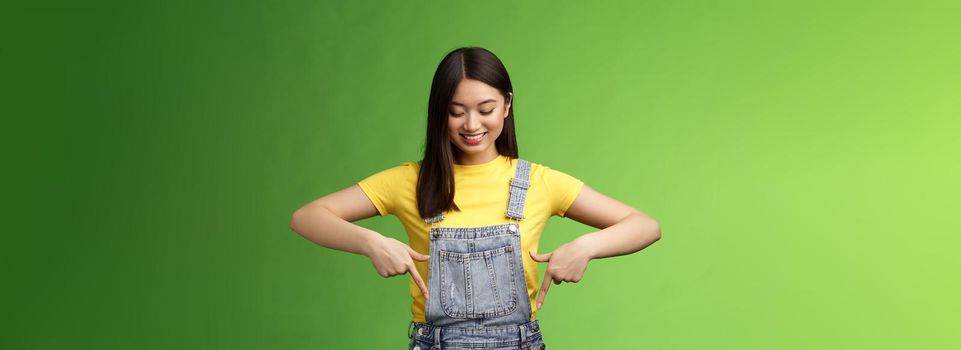 Studio shot easygoing cute asian female introduce new promo, pointing looking down amused pleasantly smiling, gladly show interesting thing, stand green background happy excited. Copy space