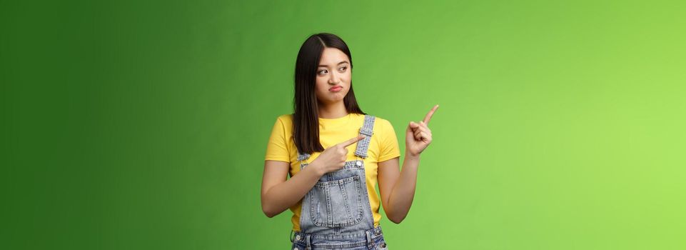 Upset cute asian female student pulling face regret shame, pity missing good chance buy product sale, pointing looking left disappointed, jealously gaze coworker, stand green background.