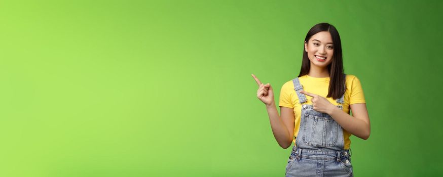 Lovely pretty tender asian woman showing way, tilt head flirty smiling, helpful pointing left, introduce plan, gladly present promo recommend cool new product, stand green background happy.