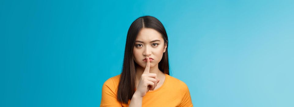 Hush stay silent. Serious-looking asian girl look displeased grumpy show shush sign press index finger lips, frowning upset, telling not speak, untell secret, prohibit loud music, blue background.