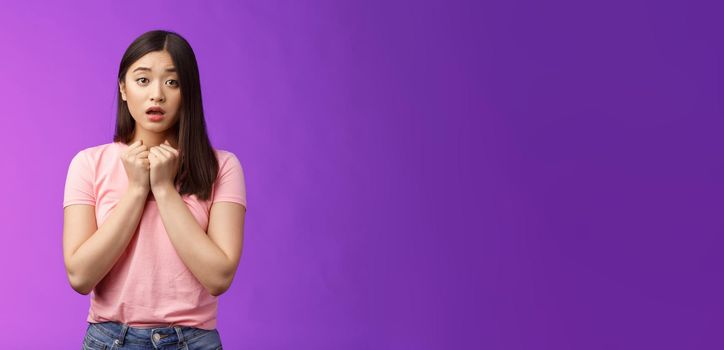 Timid concerned cute asian girl express empathy and worry, press hands heart upset, sorry for friend, open mouth, sighing sad nervous, hesitating, anxiously saying love you, stand purple background.