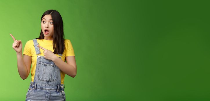 Shocked gasping alarmed pretty asian girl drop jaw impressed, turn pointing left hear loud bang, stand astonished speechless, gazing curious event, pose green background. Emotions concept
