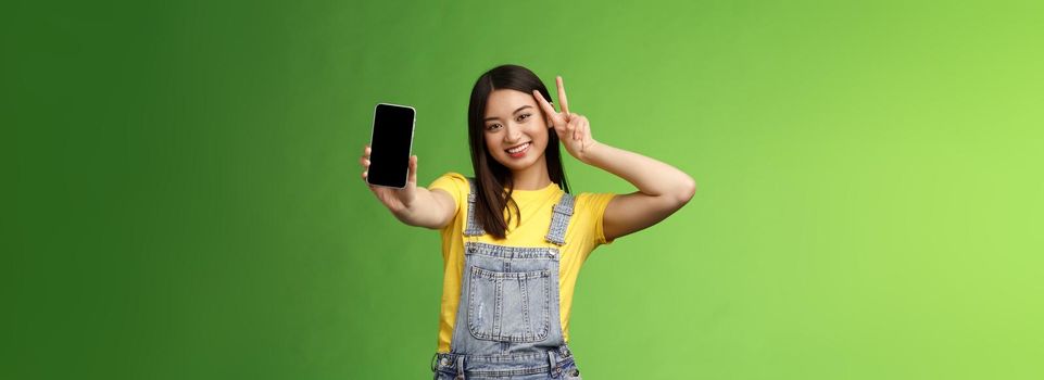 Cute carefree asian brunette showing application on smartphone screen, make victory peace sign, smiling joyfully, brag social media popularity, followers amount, stand green background.