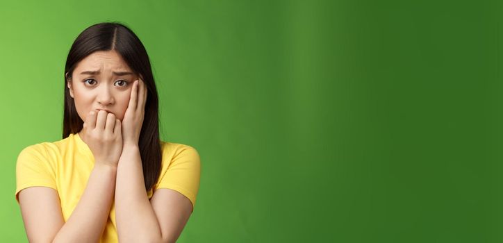 Innocent insecure timid asian scared girl panicking, standing afraid victim terrified, touch cheek shocked, frowning stunned, biting fingernails, anxiously stare camera, green background.