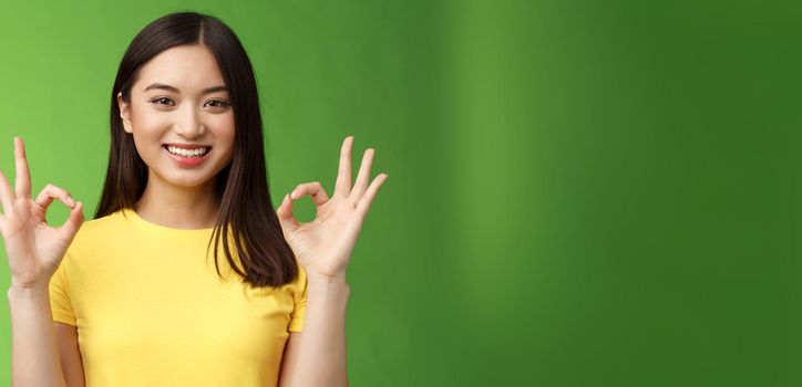 Everything under control. Friendly pleasant relaxed asian girl keep calm, assure all good, show okay ok sign, smiling approval, accept excellent idea, stand green background upbeat delighted quality.