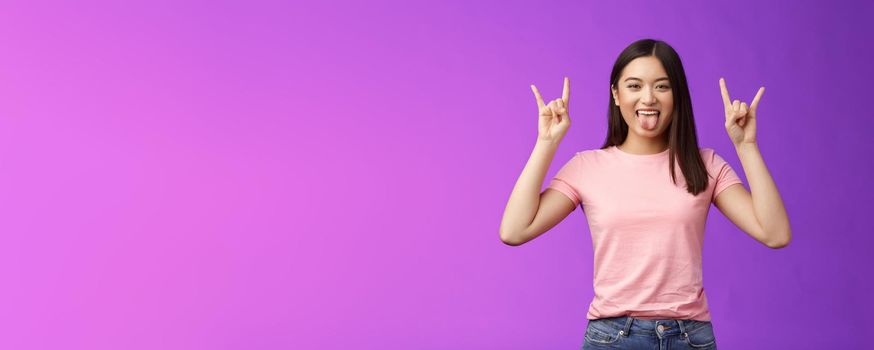 Rebellious amused asian girl enjoy rock-n-roll, attend awesome concert have fun, show tongue make heavy-metal gesture, express excitement and joy, stand purple background cheerful.