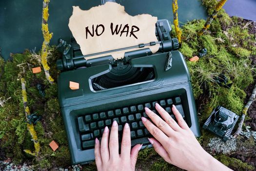 Women's hands are typing on an old retro typewriter text - no war. The field is bigger with toy tanks and soldiers. Conceptual photo. The conflict between Ukraine and Russia.