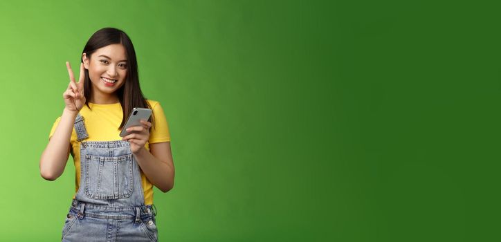 Cute tender asian woman hold smartphone, show victory peace sign, look camera delighted, carefree pose near green background, win online giveaway, triumphing joyfully beat score game.