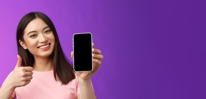 Close-up satisfied happy attractive modern asian woman give positive feedback, show smartphone display, introduce good app, raise thumb-up like application, stand purple background delighted.