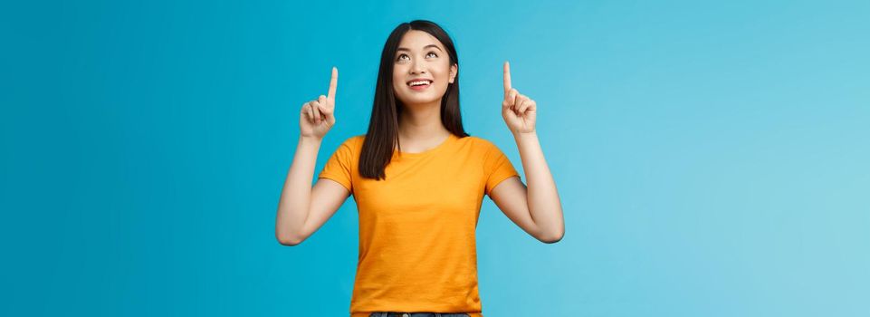 Asian girl contemplating view enjoy summer vacation like sunny days, look pointing up amused, feel enthusiastic upbeat, smiling broadly showing top promo, gaze with interest, blue background.