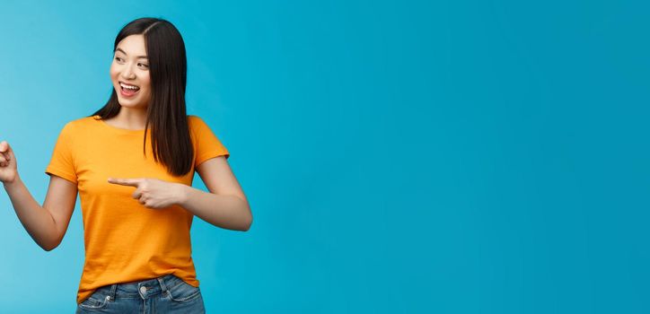 Friendly enthusiastic asian girl curiously looking sideways, pointing left interesting object, grinning joyfully discuss new opened store, stand blue background carefree lively talking.