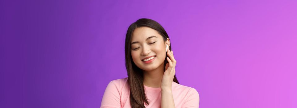 Cute asian girl enjoying listening music, put on wireless headphone, tilt head close eyes dreamy smiling satisfaction, perfect sound, make volume earphone louder, stand purple background.