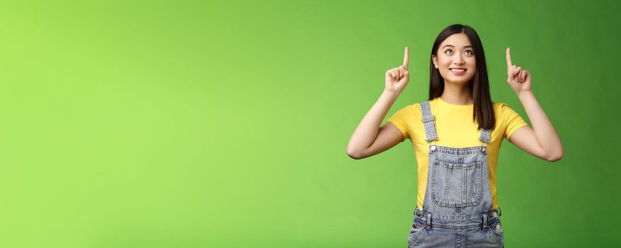 Dreamy hopeful cute asian girl look pointing up amused, smiling delighted, contemplate interesting object, enjoy stargazing, grinning satisfied, gazing top promo, stand green background.