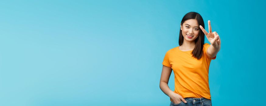 Cheerful friendly asian girl pacifist smiling happily show peace, victory sign, determined win aim achieve success stand blue background joyful, feel luck enthusiastic, wear yellow summer t-shirt.