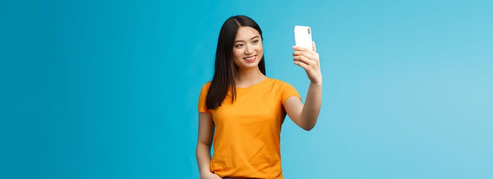 Stylish good-looking asian cute girl hold smartphone talking video-call smiling broadly look phone screen, taking selfie, feeling pretty, talking followers online social media, blue background.